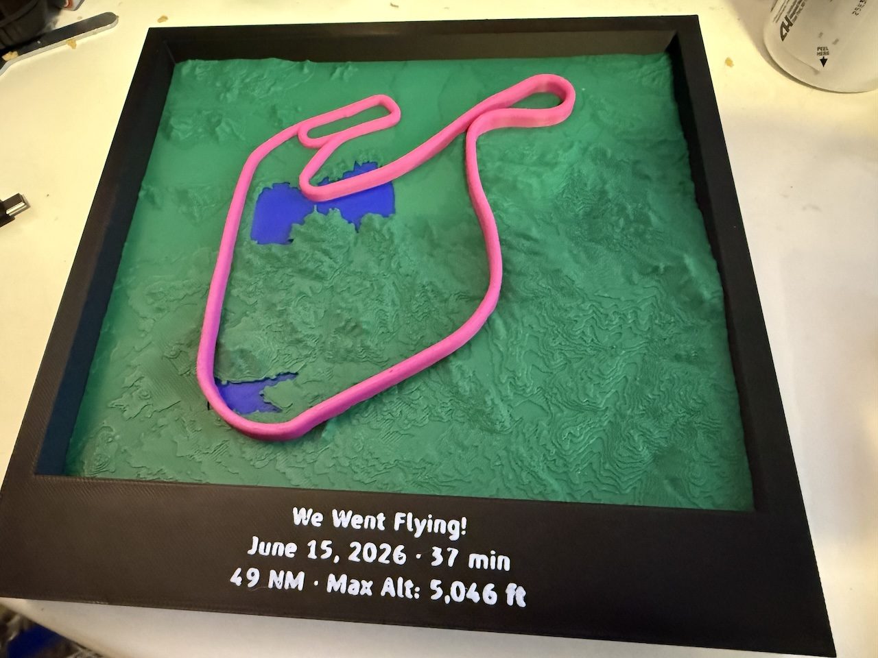 A finished print: a green relief map of the terrain in a black frame, with a pink flight path standing above it and the flight's date, duration, distance and maximum altitude on the frame below.
